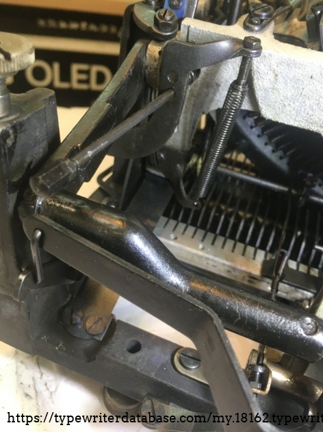 one of two springs that pull the typearm-basket against its upper stop. For capital letters the basket is lowered against a lower stop, both adjustable.
Below you see a leaf spring, being one of the links in a four-bar linkage that guides the basket up- and down movement