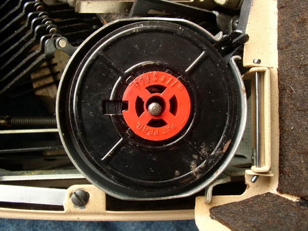 This shows the FitSall(R) spool system, which consists of a metal spool with a large center opening, probably to fit over the ribbon core of those typewriters that use cores instead of spools, and then a red plastic insert to fill that space with the more usual spool spindle. Curiously, even the hole in the insert is too big for this Smith-Corona; there might have been a yet smaller insert to take the diameter down from this size, which fits Olivettis.