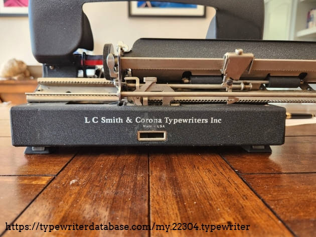 Rear view of the Smith-Corona Clipper sitting on a wooden table with the top cover open. The bottom of the black body reads: "L C Smith & Corona Typewriters Inc. Made in USA