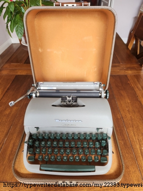 A powder coated gray typewriter with green keys sits in its open case on a wooden table.