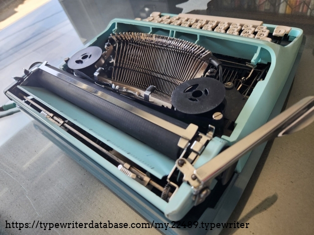 Angled view from the right hand side and behind of the Remington Streamliner typewriter with the hood removed to provide a view of the typebasket, typebars, ribbon spools and the platen.