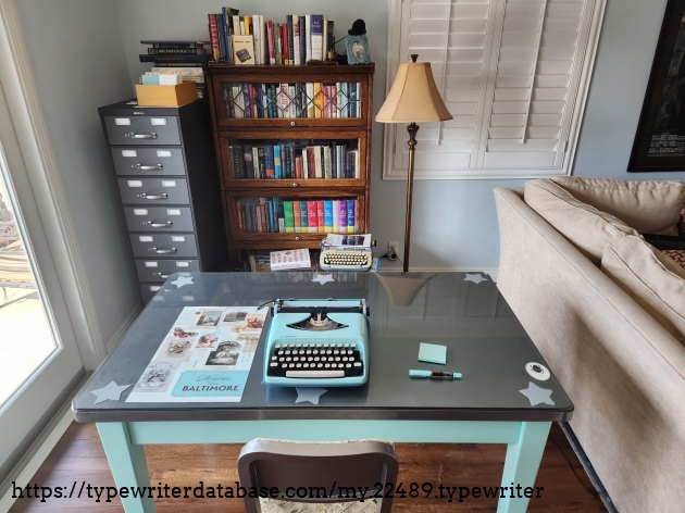 View of a working desk area featuring a silver/glass topped mint blue desk on which sits a matching colored portable typewriter, a fountain pen and a stack of Post-it notes. In the background is a card index filing cabinet and a barrister bookcase full of books.