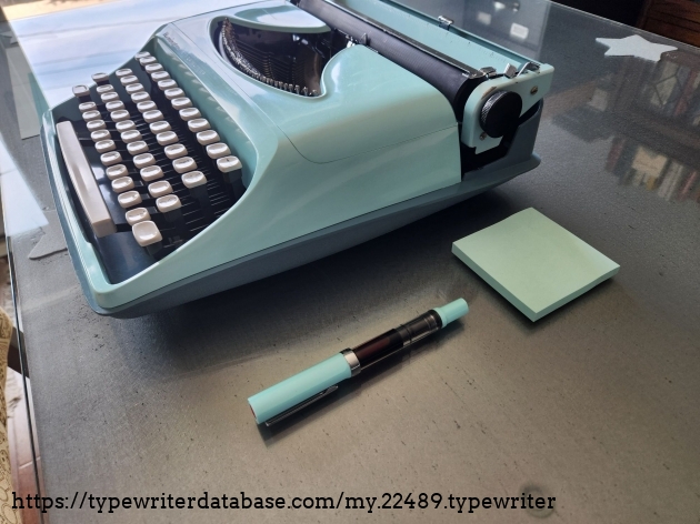 Oblique view from the right hand side of the mint blue typewriter next to which are a matching fountain pen and pad of Post-It Notes.
