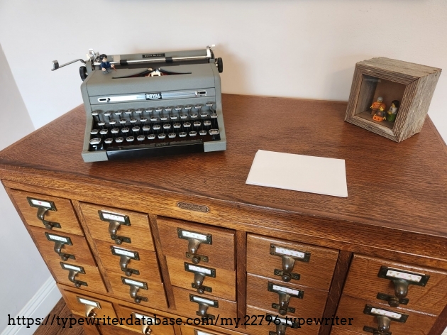 A gray typewriter sits at an angle on the top of a 20 drawer wooden library card catalog. Next to it is a stack of index cards and a small wooden shadow box with three Lego people posing inside.