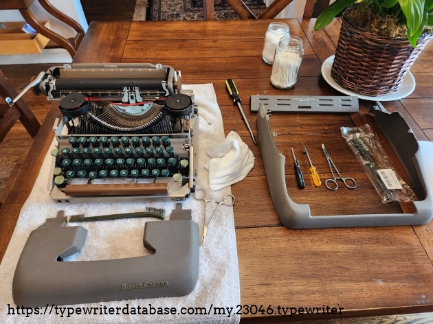 A wooden table with a towel on it. On the towel is the chassis of the typewriter with the body panels removed and sitting around it. Ordered next to the typewriter are several screwdrivers, brushes, and surgical clamps