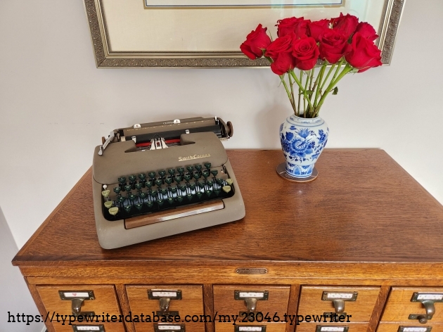 Brown bodied Smith-Corona Clipper with green keys sitting on a wooden library card catalog next to a small vase of red roses.