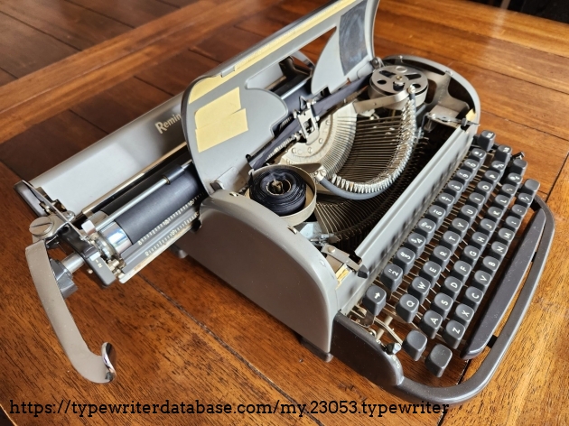 Angle down on a Remington All-New typewriter with the ribbon cover open to feature the typebasket and ribbon cups. The carriage is shifted to the far left.