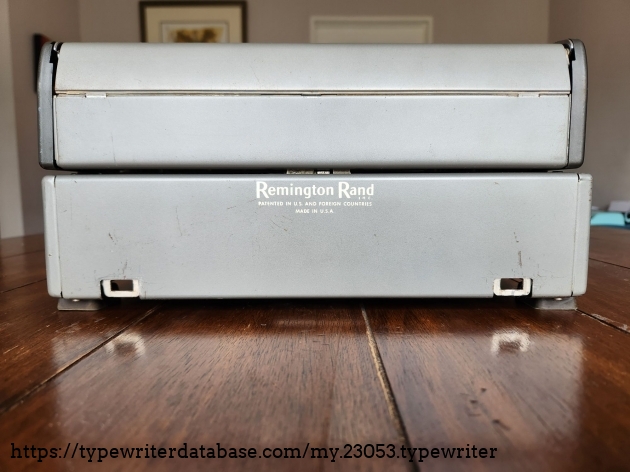 Straight on view of the rear of a Remington All-New typewriter. On the bottom half is a logo that reads Remington Rand Inc. Patented in the US and Foreign Countries Made in the U.S.A. At the bottom of the machine are two open metal slots on opposite sides into which metal tabs on the case would be used to stabilize the machine for carrying.