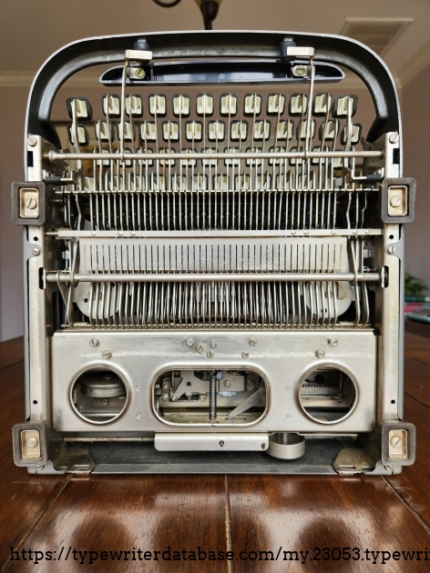 View of the bottom of a Remington All-New typewriter. We see the bottom of the keys and the many dozens of thin metal bars connecting the keys to the typebars. At the bottom is a metal plate with a variety of adjustment screws for properly aligning the typewriter. We can just make out the round shape of the bell at the bottom of the machine. There are four rectangular rubber feet at the corners of the unit.