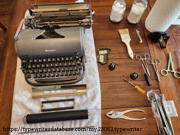 Remington All-New typewriter laid out on on a towel on a wooden table. The platen knobs and platen have been removed. The paper pan, rollers and platen are laid out in front of the typewriter and spread around the typewriter are a variety of screwdrivers, pliers, and other tools.