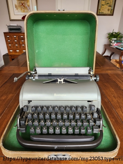 Opened typewriter case with a green interior. Sitting inside it is a rounded Remington All-New typewriter in gunmetal gray with dark gray keys. The two are sitting on a rustic wooden dining room table with a library card catalog just visible in the background.