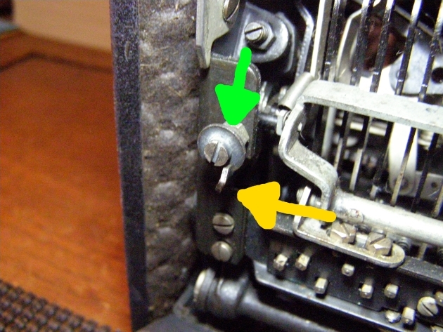 Yellow arrow points toward a catch attached to the carriage which is pushed back when the shift key is pressed, the green arrow points to a disc the catch hooks onto.  This keeps the carriage from bobbing with the vibrations in a ship or an airplane.