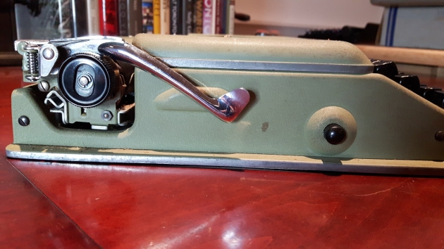 The return lever folds down and hooks under the left platen knob. The black circle on the right is a button that pushes in and pops back out through a hole in the cover, holding it in place on either side of the typewriter.